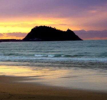 Atardecer en la playa de Getaria con colores cálidos reflejados en el agua y el icónico monte San Antón al fondo.